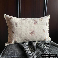A decorative pillow with a floral insect pattern, featuring a beige color with detailed insect designs, sitting on a dark wood background.