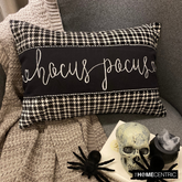 Black and white applique houndstooth lumbar pillow cover with embroidered 'Hocus Pocus' text on the center in cursive, placed on a gray couch and a gray throw with a skull and book in the foreground.
