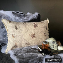A pillow with a floral insect design on a beige fabric, surrounded by Halloween-themed decorations such as a skull, books, and a pumpkin, set against a webbed backdrop.
