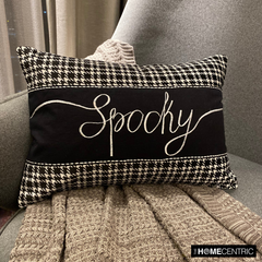 Halloween-inspired throw pillow with the word 'Spooky' written in a white, elegant script, set against a black and white houndstooth pattern, perfect for Halloween home decor, set on a grey textured throw on a gray chair.