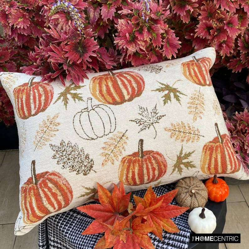 Orange, beige & ivory color pumpkin pattern with maple leaves lumbar pillow cover crafted on a woven jacquard fabric for fall & Halloween decor from The HomeCentric's autumn decor collection. The oblong cushion cover is set on a black stool covered with a houndstooth pattern throw, paired ith maple leaves and white, orange, & burlap pumpkins, to create a cozy fall season vibe, in a natural setting with orange-red leaves in the background. Explore more of The HomeCentrc's Autumn Decor.
