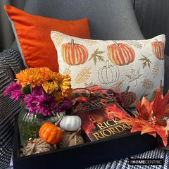 Orange, beige & ivory color pumpkin pattern with maple leaves lumbar pillow cover crafted on a woven jacquard fabric for fall & Halloween decor from The HomeCentric's autumn decor collection. The oblong cushion cover is set on a grey chair covered with a black & white checks throw, and layered with an orange solid velvet throw pillow cover. A finishing touch is added with a tray containing maple leaves, book, burlap pumpkins and flowers in orange and purple color in a vase.