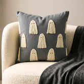 Halloween themed skeleton holding a grey decorative embroidered throw pillow cover with cute ghost design placed on a black chair covered with spider web, decorated with a skull and a witch pot kept on a black book to create a Halloween look.