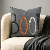 Halloween decoration with a skeleton holding a 'BOO!' charcoal embroidered throw pillow cover surrounded by spooky props such as a skull, pumpkins, and witch pot.