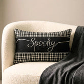 Halloween-inspired throw pillow with the word 'Spooky' written in a white, elegant script, set against a black and white houndstooth pattern, perfect for Halloween home decor. Set on a grey chair with a textured grey throw, surrounded by Halloween decor such as a skull, witch pot with white smoke, and a spider.