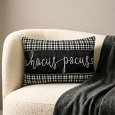 Black and white applique houndstooth lumbar pillow cover with embroidered 'Hocus Pocus' text on the center in cursive, placed on a gray couch and a gray throw with a skull and book in the foreground.