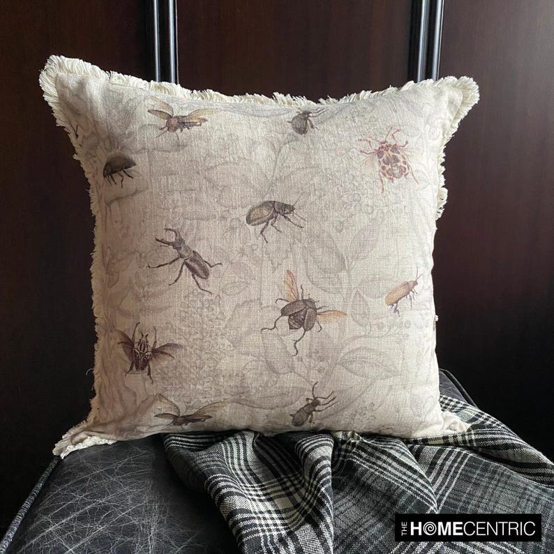 Close-up of a decorative pillow featuring a vintage insect design with bees, beetles, and flies on a neutral-colored linen fabric with fringe detailing, settled on a black stool with the checkered black & white throw against a wooden background.