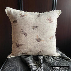 Close-up of a decorative pillow featuring a vintage insect design with bees, beetles, and flies on a neutral-colored linen fabric with fringe detailing, settled on a black stool with the checkered black & white throw against a wooden background.