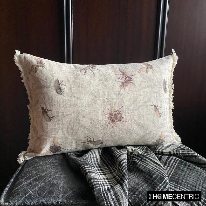 A decorative pillow with a floral insect pattern, featuring a beige color with detailed insect designs, sitting on a dark wood background.