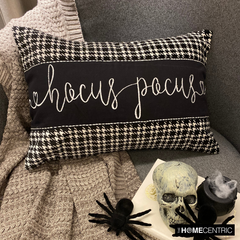 Black and white applique houndstooth lumbar pillow cover with embroidered 'Hocus Pocus' text on the center in cursive, placed on a gray couch and a gray throw with a skull and book in the foreground.