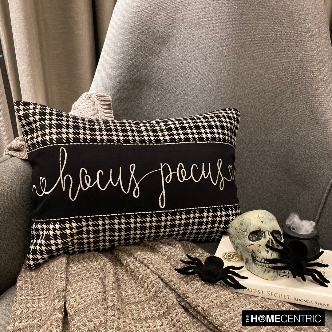 Halloween movie-themed pillow cover with 'Hocus Pocus' text in white cursive, featuring a houndstooth pattern, placed with skull and spider decorations on a cozy gray throw on a gray chair.