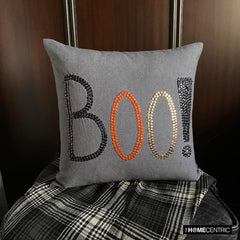 Halloween-themed throw pillow cover with the word 'BOO!' in black, orange, and gold sequins, placed on a black and white monochrome throw against a wooden background. 