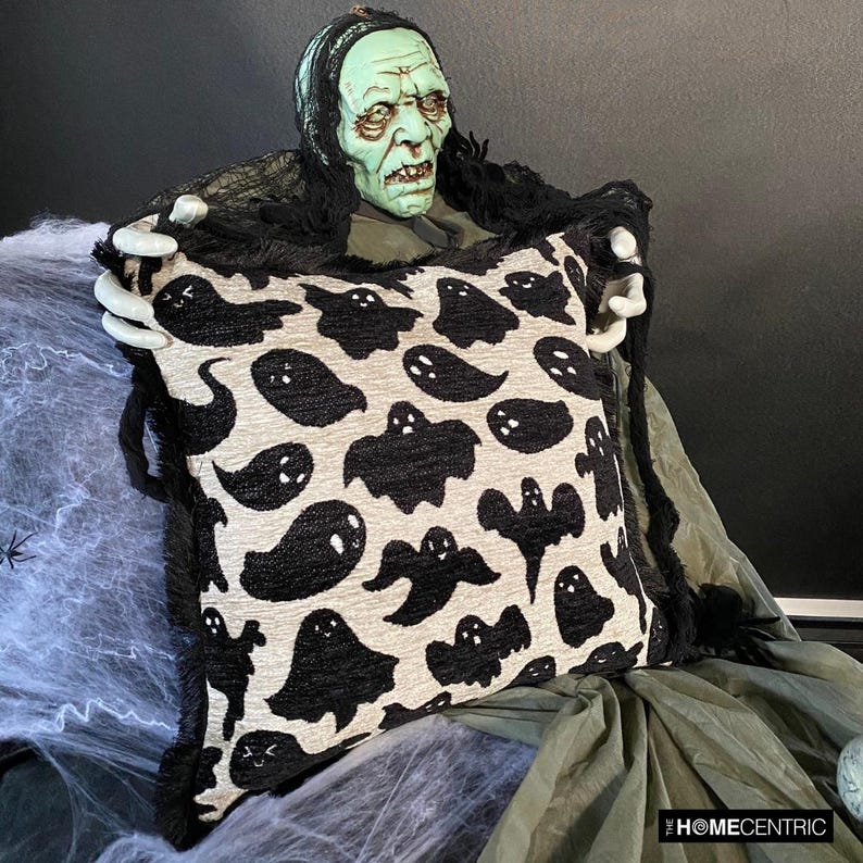 Close-up of ghost-themed Halloween pillow with black fringe held by a scary Halloween ghost/skeleton placed on a black chair covered with spider web.