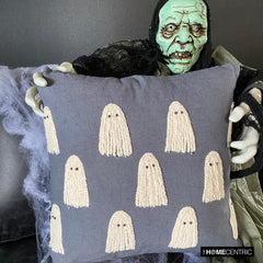 Close-up of Halloween scary skeleton holding an embroidered ghost cushion cover in grey color placed on a chair covered with spider web. 