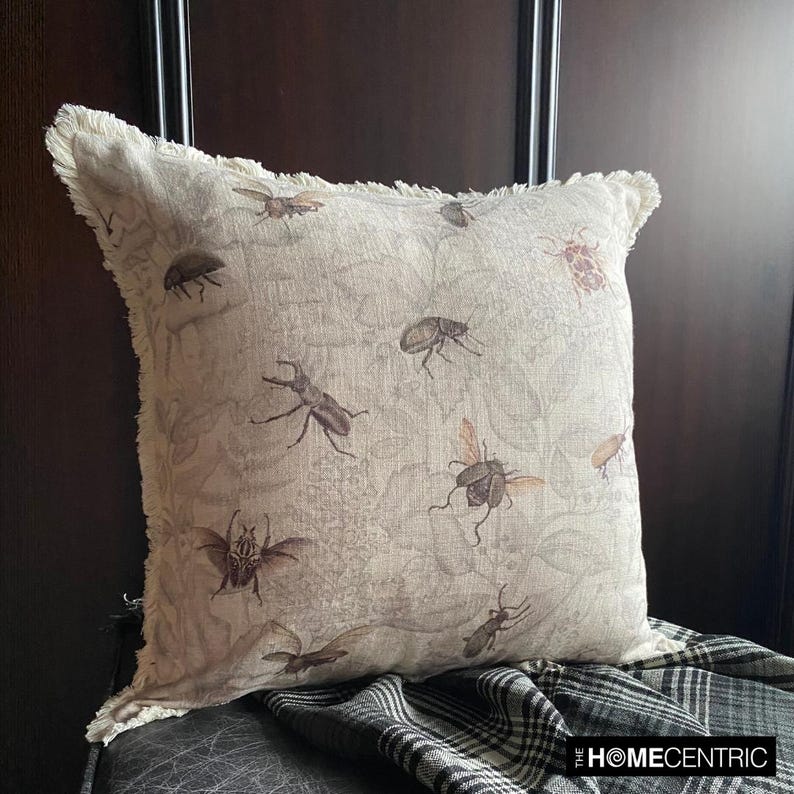 Close-up of a decorative pillow with an intricate vintage insect pattern, including beetles and bees, on a neutral linen fabric with frayed edges.