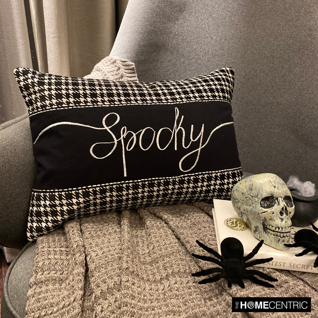 A Halloween throw pillow featuring 'Spooky' in script with black and white houndstooth borders, complemented by a skull and spider for a spooky touch, set on a grey chair with textured grey throw.