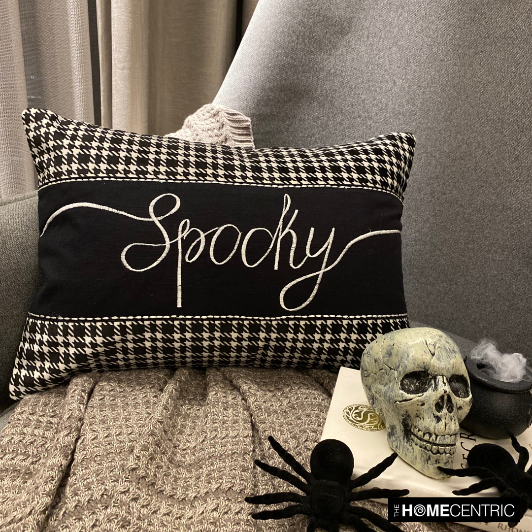 Elegant 'Spooky' Halloween pillow with black and white houndstooth details, accented by a skull, spiders, witch pot with white xmoke and cozy autumn decor for a festive vibe. Set on a grey chair and paired with a gray textured throw. 