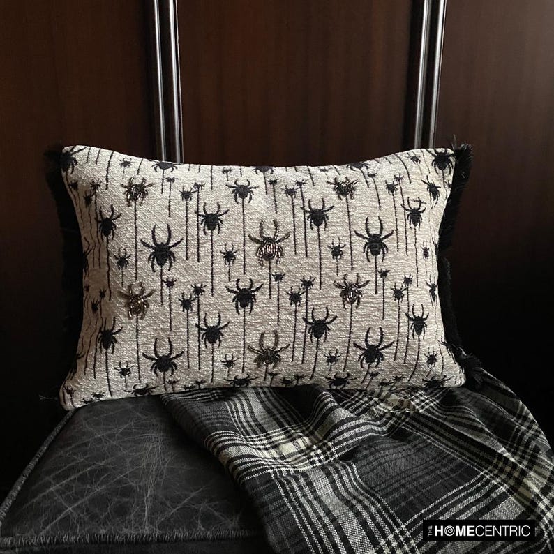 Halloween lumbar pillow cover with spider design on a woven jacquard fabric and beads and lace embroidery, set against a dark wood background with a plaid blanket.