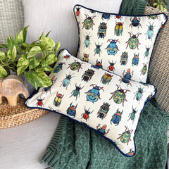 Two decorative pillow covers with vibrant beetle and bug embroidered prints, placed on a green knitted throw and surrounded by plants.
