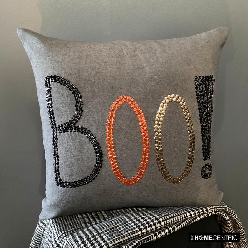 Halloween pillow cover with 'BOO!' embroidered in black, orange, and gold sequins on grey fabric, placed on a black stool with a checkered throw.