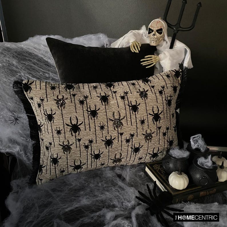 Halloween pillow with black spider silver beads embroidery, skeleton figure, and Halloween-themed decor including small pumpkins, witch pot with white smoke, spiders and books.