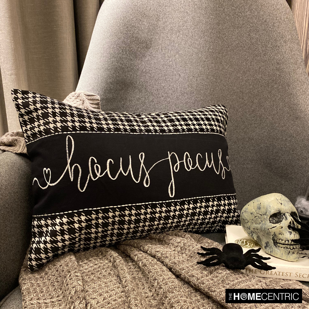 Hocus Pocus themed cotton, houndstooth patterned pillow cover with cursive white lettering, set against a gray chair and a gray throw, surrounded by Halloween-themed decor like a skull and spiders.