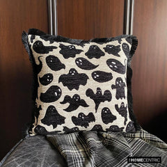 Halloween-themed pillow cover featuring ghost print on neutral fabric, set on a black chair with a checkered black & white throw against a brown wood background.