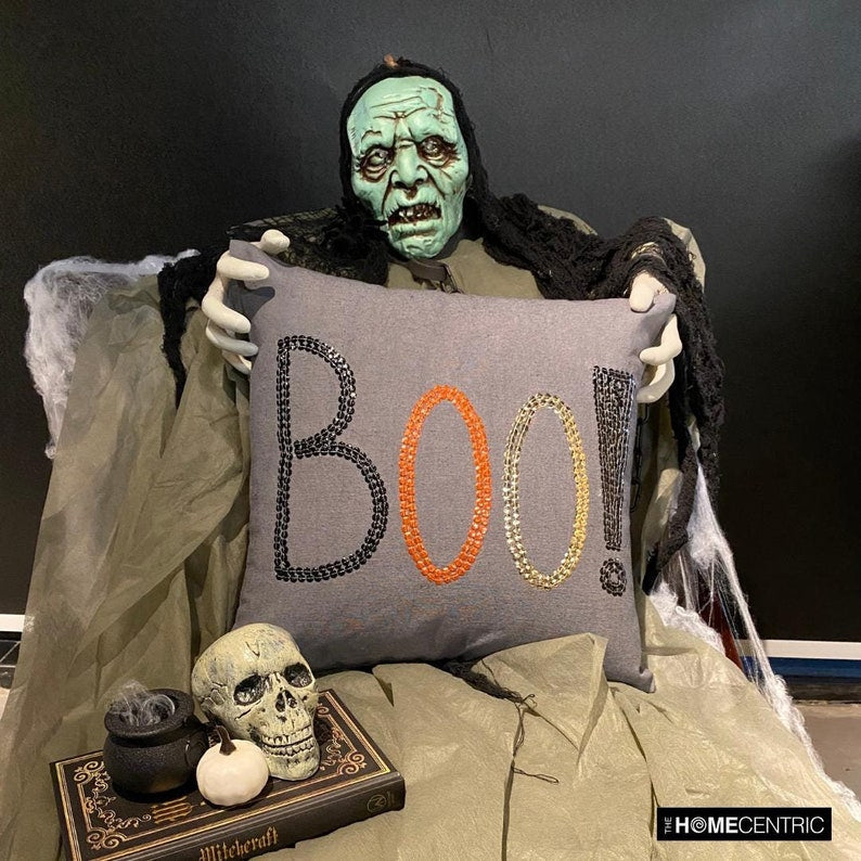 Halloween decoration with a skeleton holding a 'BOO!' charcoal embroidered throw pillow cover surrounded by spooky props such as a skull, pumpkins, and witch pot.