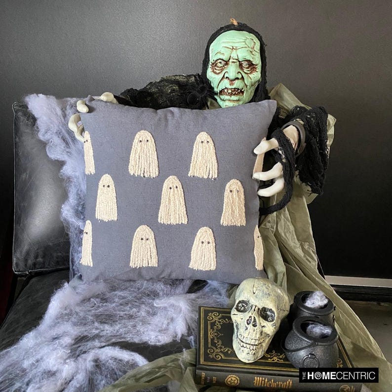 Halloween themed skeleton holding a grey decorative embroidered throw pillow cover with cute ghost design placed on a black chair covered with spider web, decorated with a skull and a witch pot kept on a black book to create a Halloween look. 