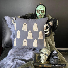 Halloween themed skeleton holding a grey decorative embroidered throw pillow cover with cute ghost design placed on a black chair covered with spider web, decorated with a skull and a witch pot kept on a black book to create a Halloween look. 