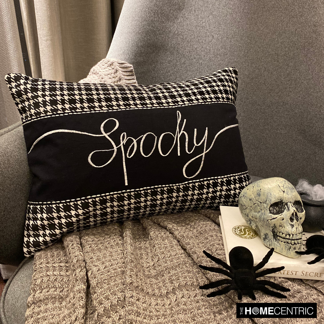 Stylish Halloween-themed throw pillow with the word 'Spooky' in white cursive on a black fabric with houndstooth pattern accents, paired with a skull, spider, a witch pot with white flames on a book, placed on a grey chair with a textured grey throw.