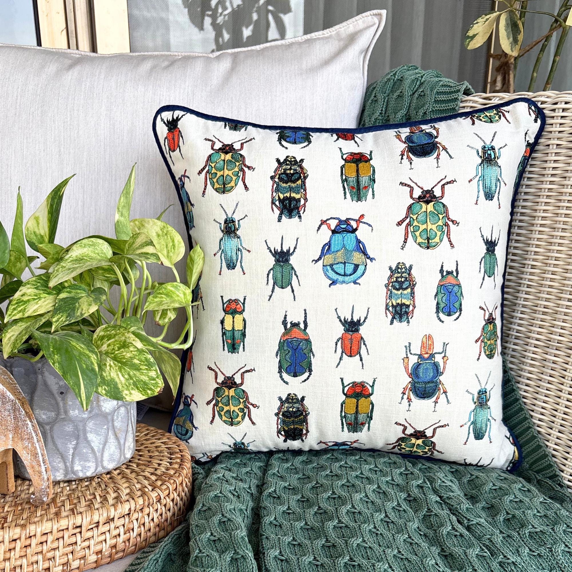 Colorful beetle-patterned embroidered throw pillow cover, surrounded by a textured green throw and a plant in an outdoor setting
