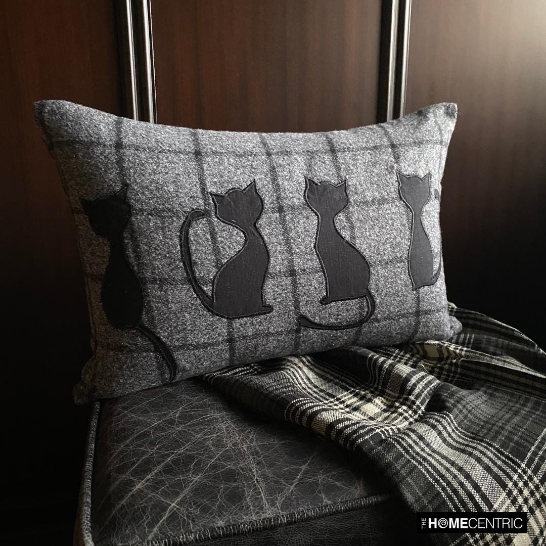 Close-up of a black cat-themed decorative pillow on wool plaid fabric, ideal for Halloween or fall decor, on a black & white plaid throw against a wooden background.