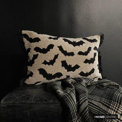 A Halloween-themed display featuring a decorative pillow with a gothic bat pattern on a black stool paired with a black & white throw, surrounded by a black background. 