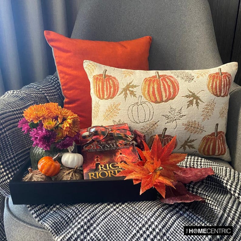 Orange, beige & ivory color pumpkin pattern with maple leaves lumbar pillow cover crafted on a woven jacquard fabric for fall & Halloween decor from The HomeCentric's autumn decor collection. The oblong cushion cover is set on a grey chair covered with a black & white checks throw, and layered with an orange solid velvet throw pillow cover. A finishing touch is added with a tray containing maple leaves, book, burlap pumpkins and flowers in orange and purple color in a vase. 