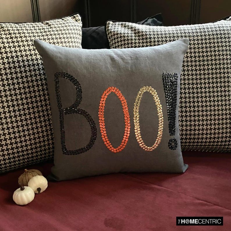Halloween pillow with a 'BOO!' design in black, orange, and gold sequins on a grey fabric, on a marron couch paired with black & white checkered euro sham.