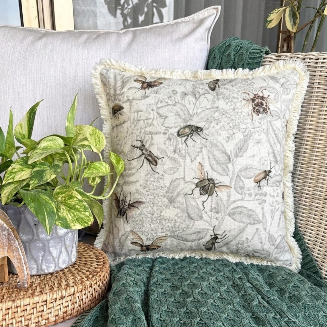 Close-up of a decorative pillow featuring a vintage insect design with bees, beetles, and flies on a neutral-colored linen fabric with fringe detailing, settled on a black stool with the checkered black & white throw against a wooden background.