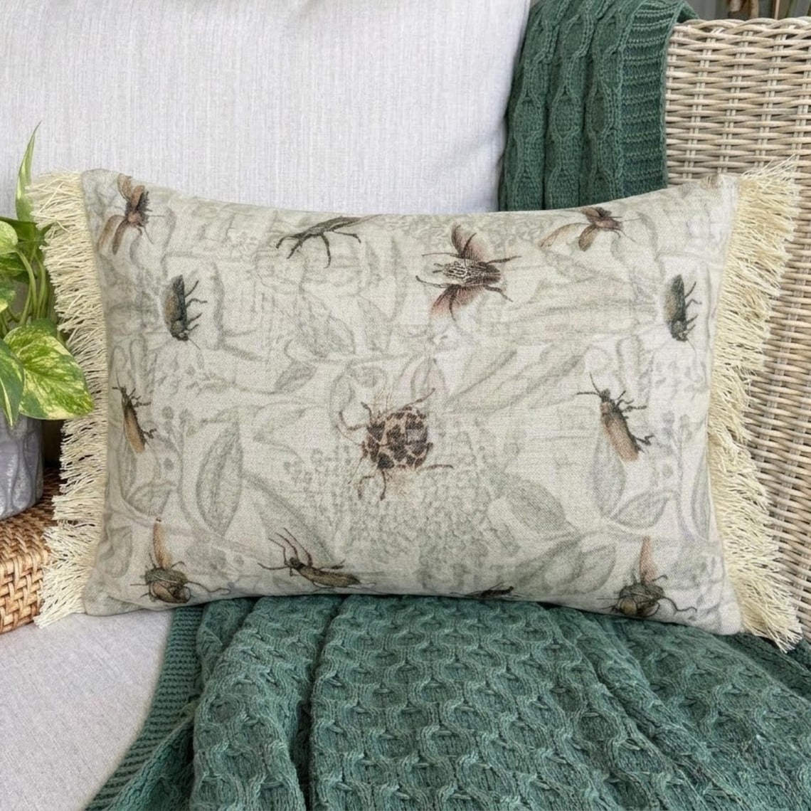 A decorative pillow with a floral insect pattern, featuring a beige color with detailed insect designs, sitting on a dark wood background.