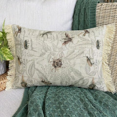 A decorative pillow with a floral insect pattern, featuring a beige color with detailed insect designs, sitting on a dark wood background.