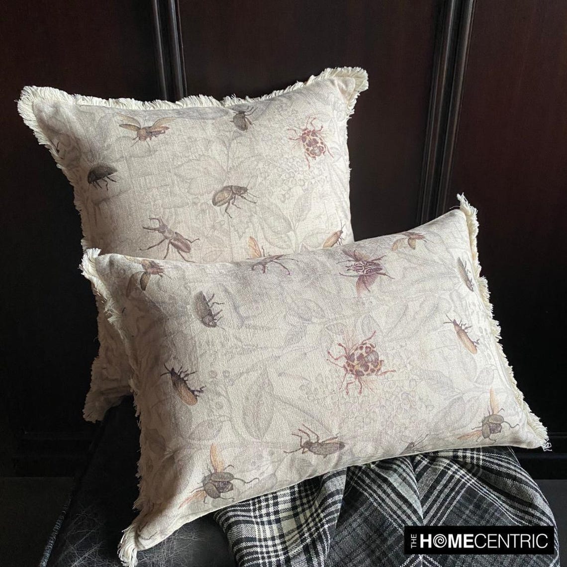 Two decorative pillows with an insect pattern on beige fabric, displayed against a dark background with a checkered cloth underneath.
