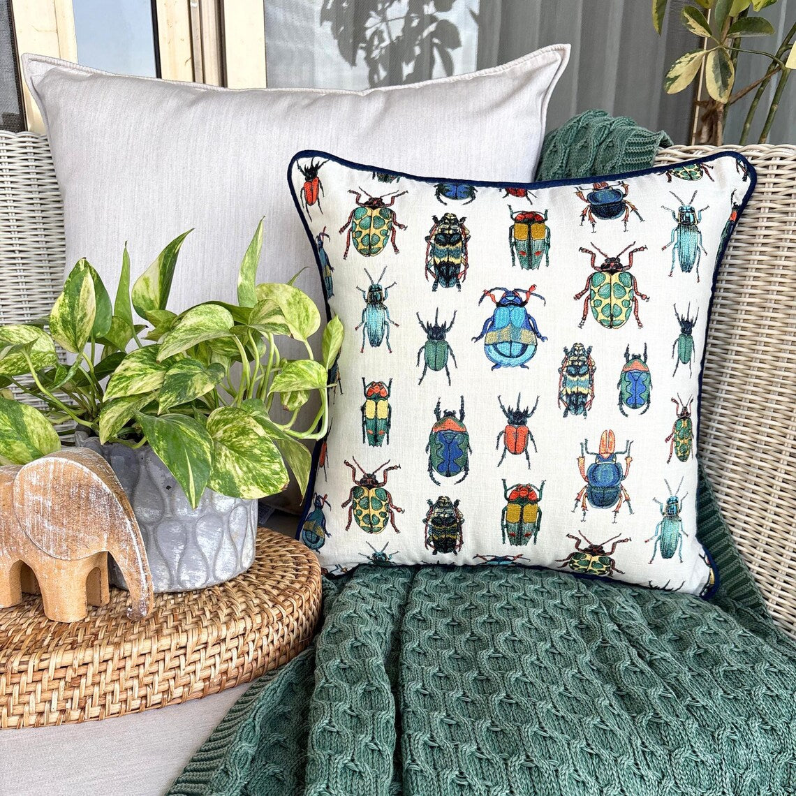 Colorful beetle-patterned embroidered throw pillow cover, surrounded by a textured green throw and a plant in an outdoor setting