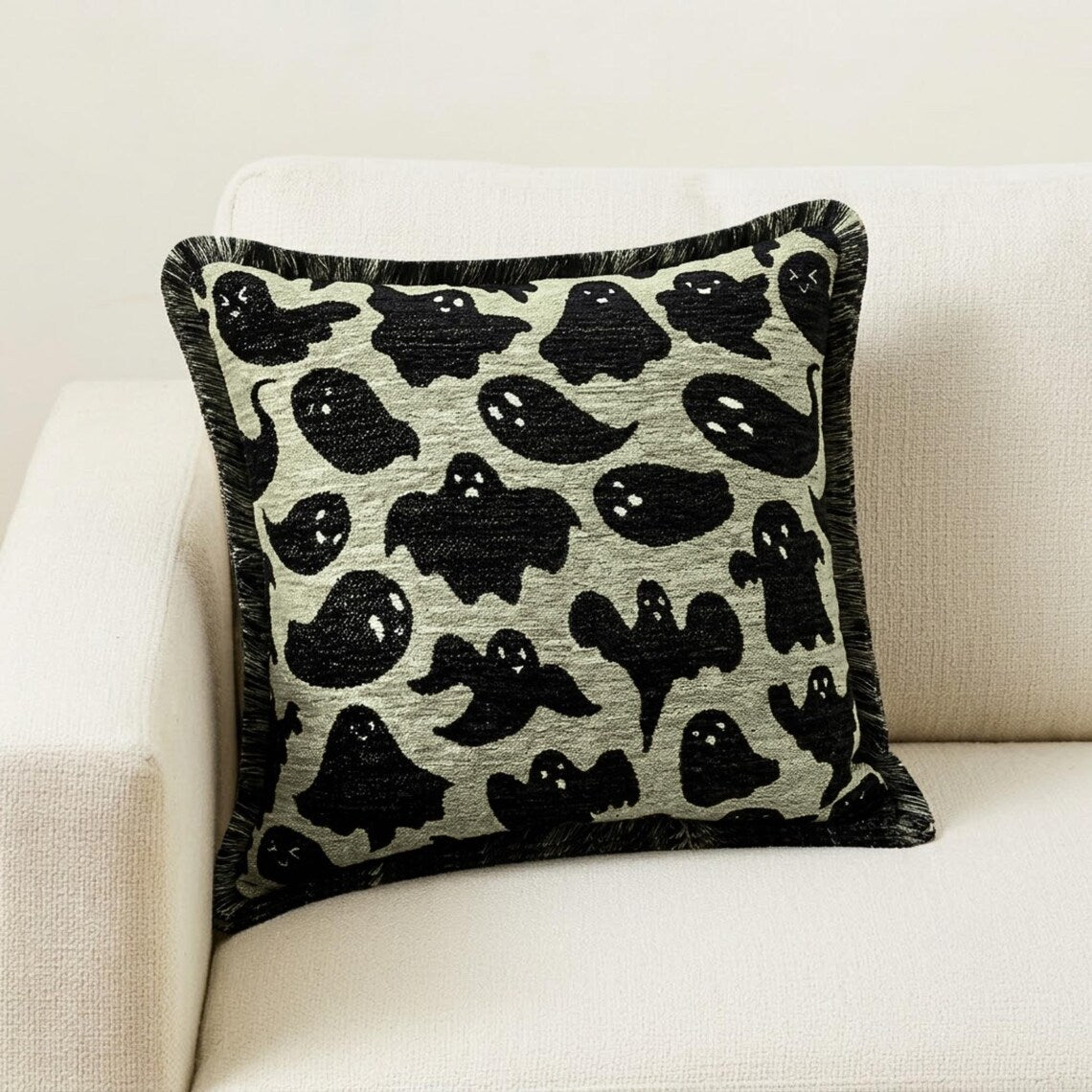 Halloween-themed pillow cover featuring ghost print on neutral fabric, set on a black chair with a checkered black & white throw against a brown wood background.