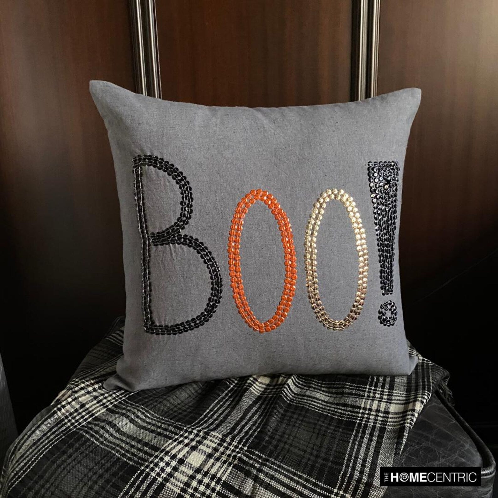 Halloween pillow with a 'BOO!' design in black, orange, and gold sequins on a grey fabric, on a marron couch paired with black & white checkered euro sham.