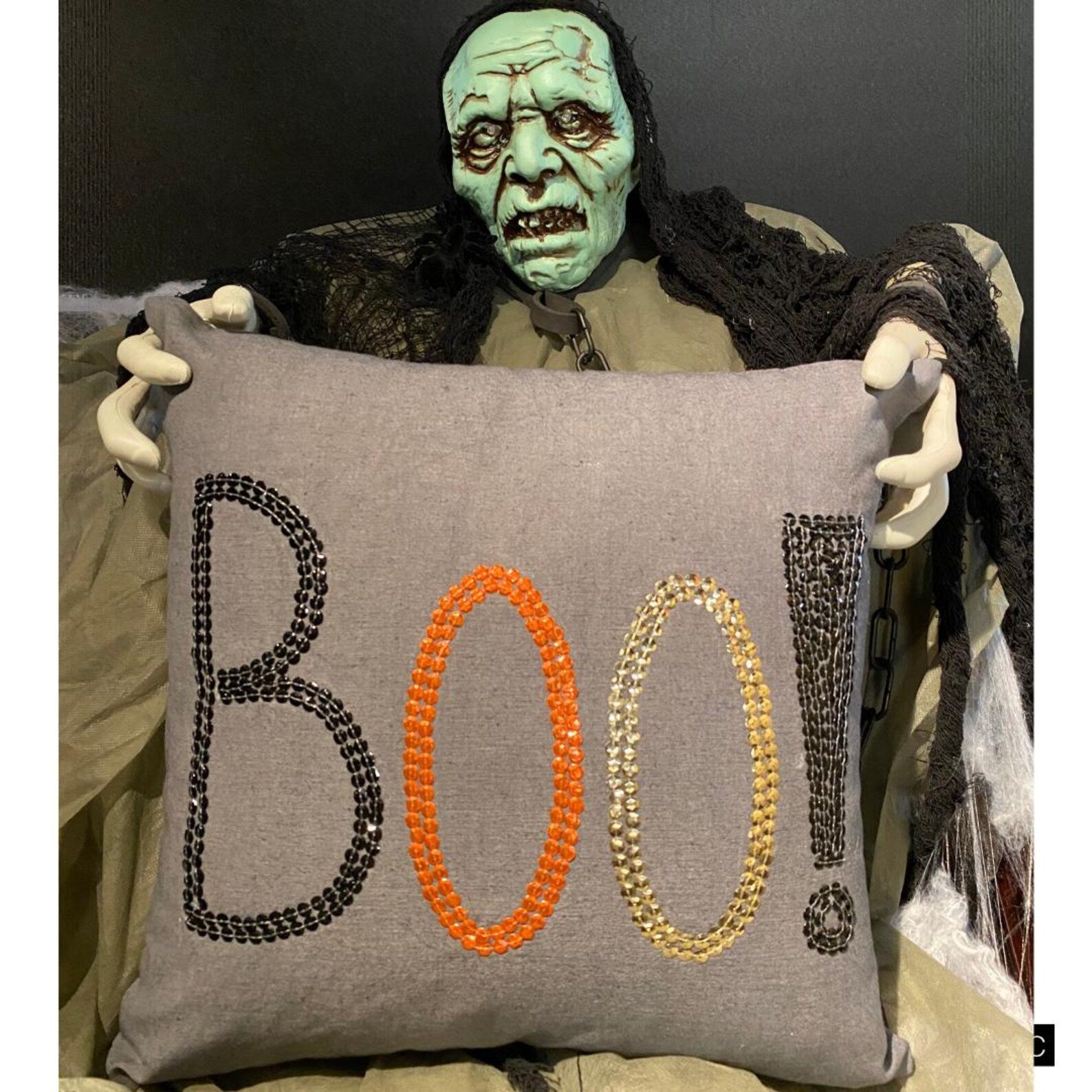 Halloween pillow cover with 'BOO!' embroidered in black, orange, and gold sequins on grey fabric, placed on a black stool with a checkered throw.
