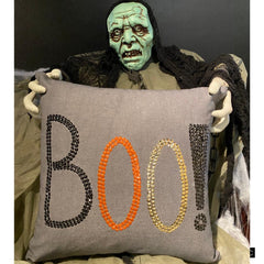 Halloween pillow cover with 'BOO!' embroidered in black, orange, and gold sequins on grey fabric, placed on a black stool with a checkered throw.