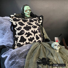 Close-up of ghost-themed Halloween pillow with black fringe held by a scary Halloween ghost/skeleton placed on a black chair covered with spider web.