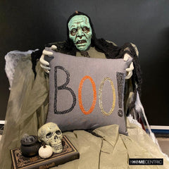 Close-up of a Halloween pillow with 'BOO!' embroidered design in black, orange, and gold sequins on a grey linen fabric held by a scary Halloween ghost.