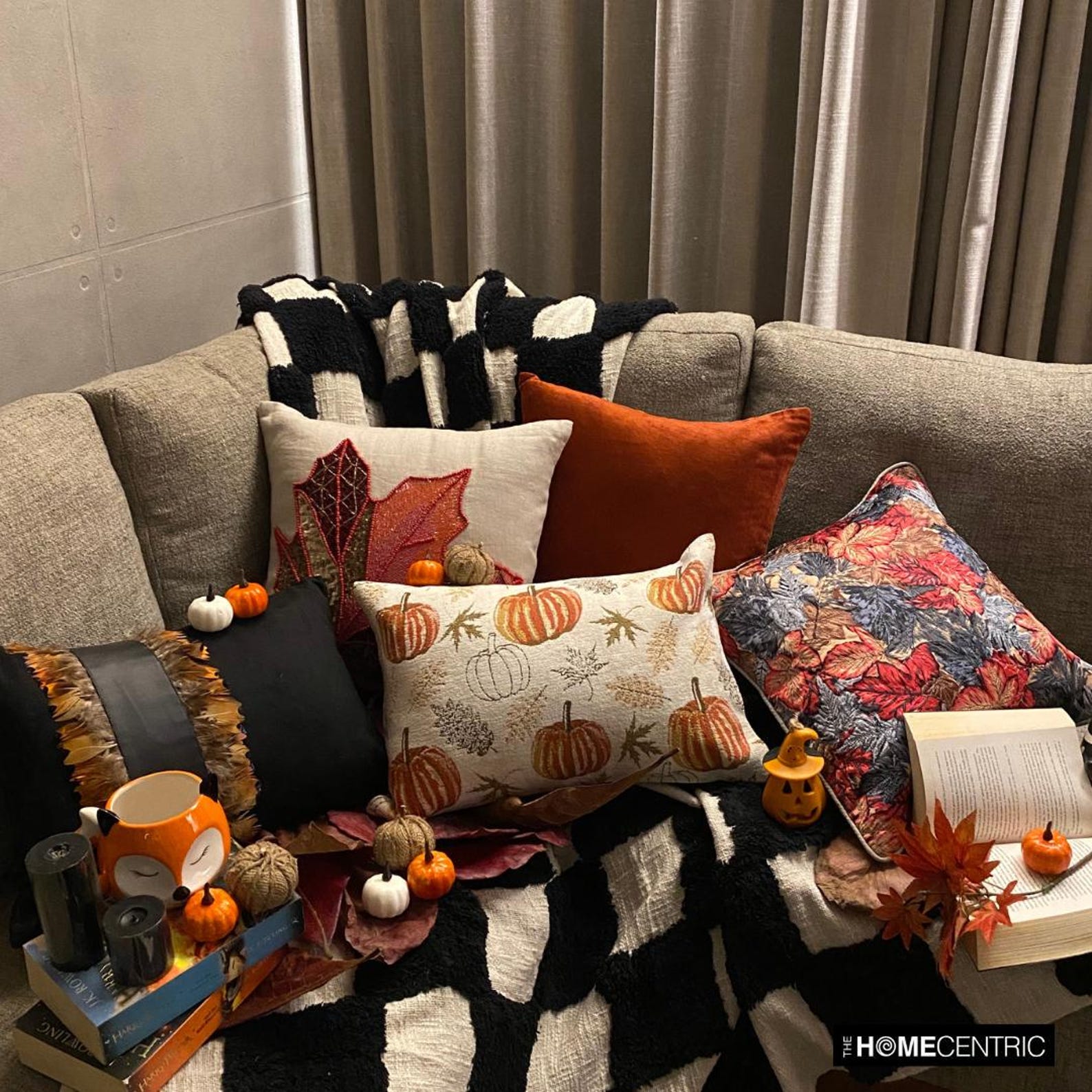 Orange, beige & ivory color pumpkin pattern with maple leaves lumbar pillow cover crafted on a woven jacquard fabric for fall & Halloween decor from The HomeCentric's autumn decor collection. The oblong cushion cover is set on a black stool covered with a houndstooth pattern throw, paired ith maple leaves and white, orange, & burlap pumpkins, to create a cozy fall season vibe, in a natural setting with orange-red leaves in the background. Explore more of The HomeCentrc's Autumn Decor.
