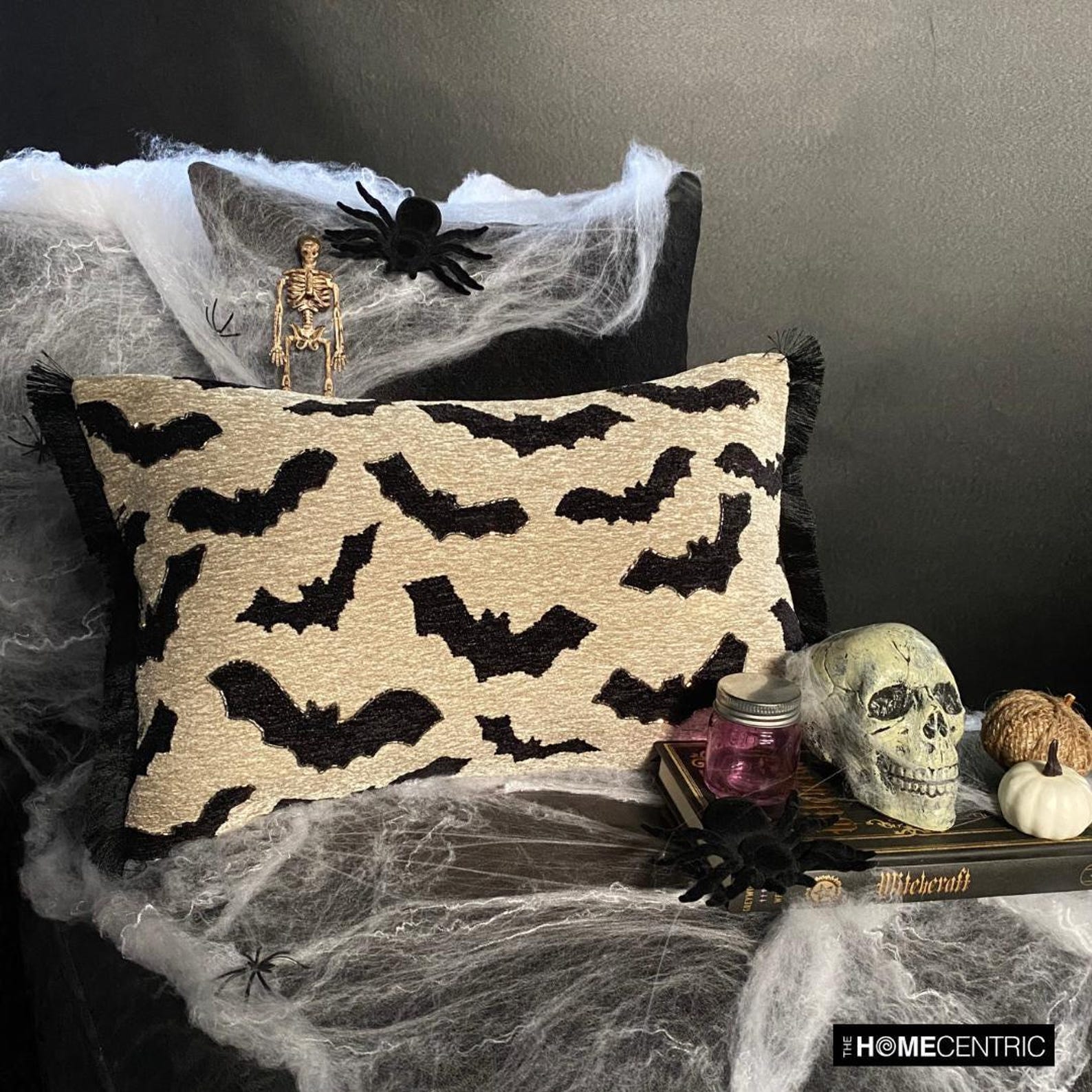 A Halloween-themed display featuring a decorative pillow with a gothic bat pattern on a black stool paired with a black & white throw, surrounded by a black background. 