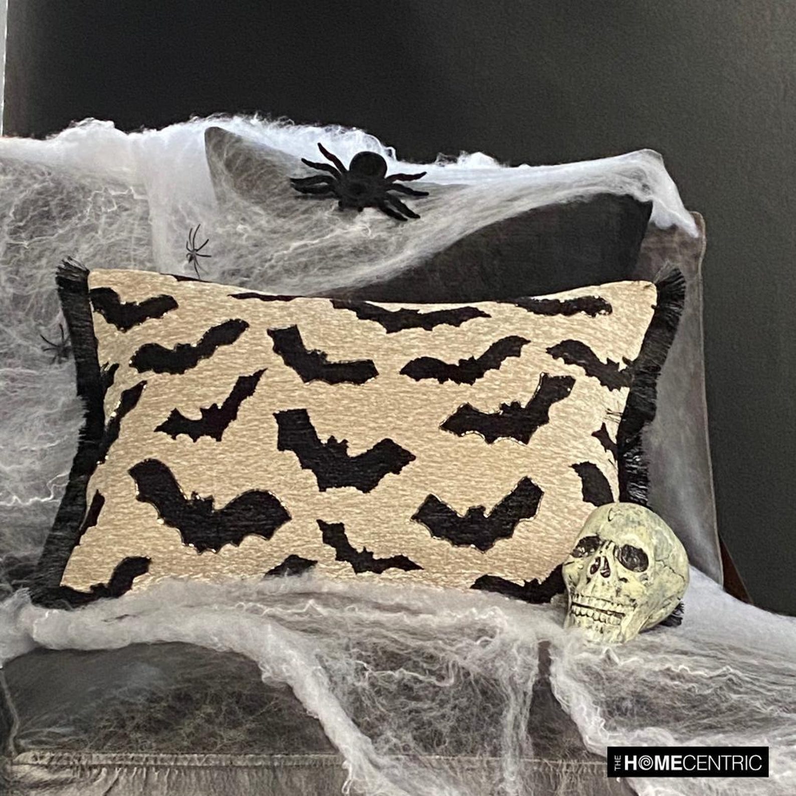Halloween-themed decor featuring a black and gray pillow with a bat print resting in a decorative cauldron, surrounded by spider web accents and a few spiders on the wall.